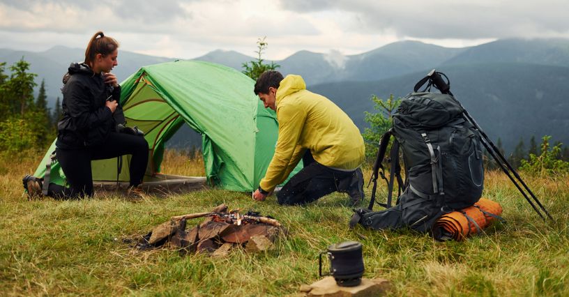 Para w odzieży trekkingowej rozbija namiot na górskiej polanie, mają ze sobą plecaki turystyczne, przygotowane drewno na ognisko i kubek turystyczny