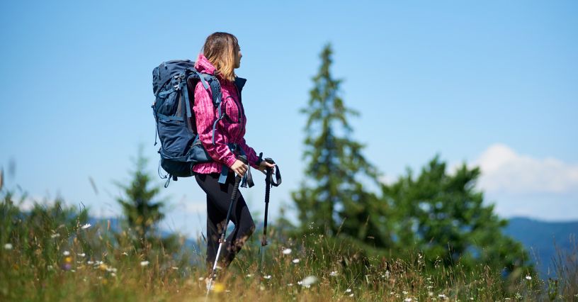Kobieta z plecakiem turystycznym i w kurtce przeciwdeszczowej idzie górską łąką używając kijków trekkingowych