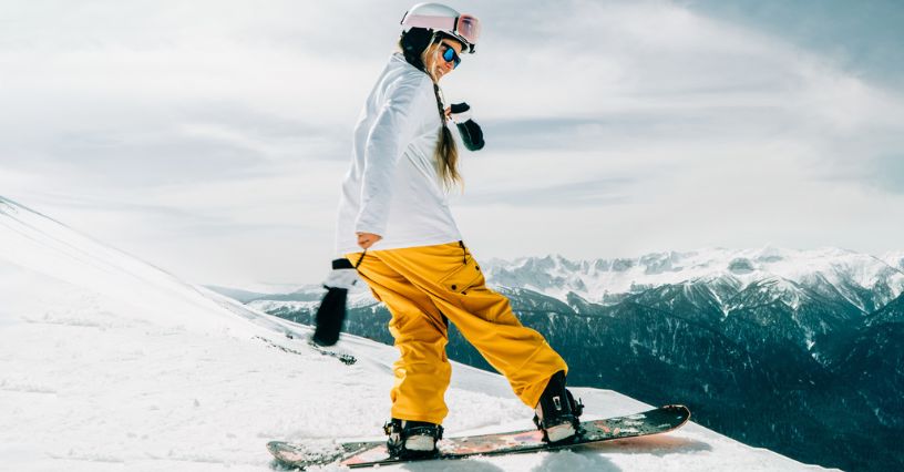 Kobieta w odzieży snowboardowej i goglach zjeżdża po stoku na desce snowboardowej