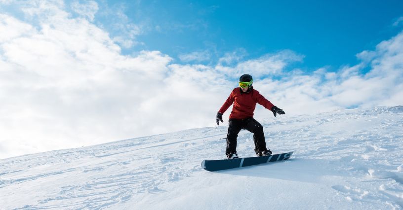 Mężczyzna w odzieży snowboardowej i goglach zjeżdża po stoku na desce snowboardowej