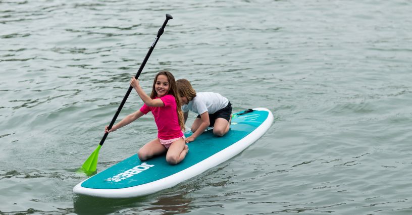 Dziewczynka z wiosłem i chłopiec pływający na siedząco na desce SUP na spokojnej wodzie