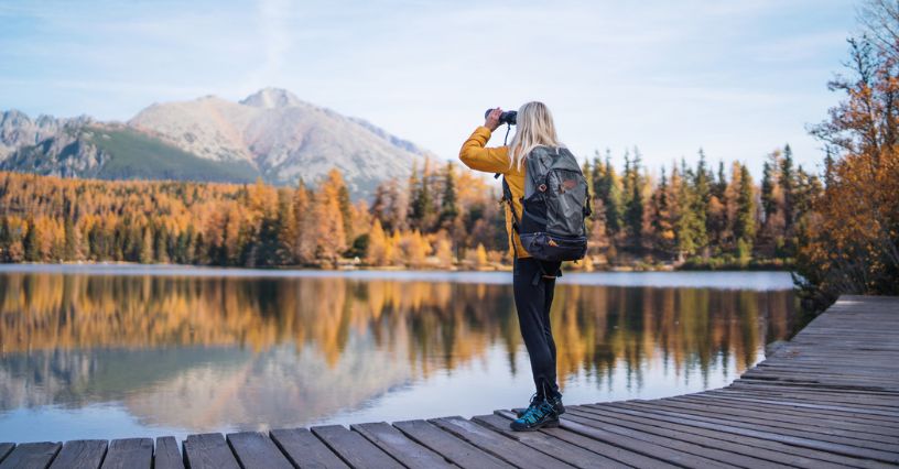 Kobieta stojąca na drewnianym pomoście nad jeziorem oglądająca przez lornetkę góry jesienią