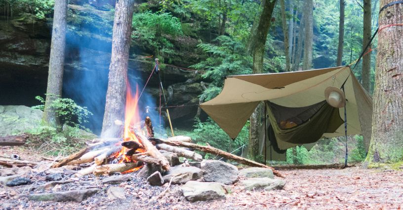 Mężczyzna leżący w hamaku turystycznym osłoniętym tarpem przy ognisku w lesie