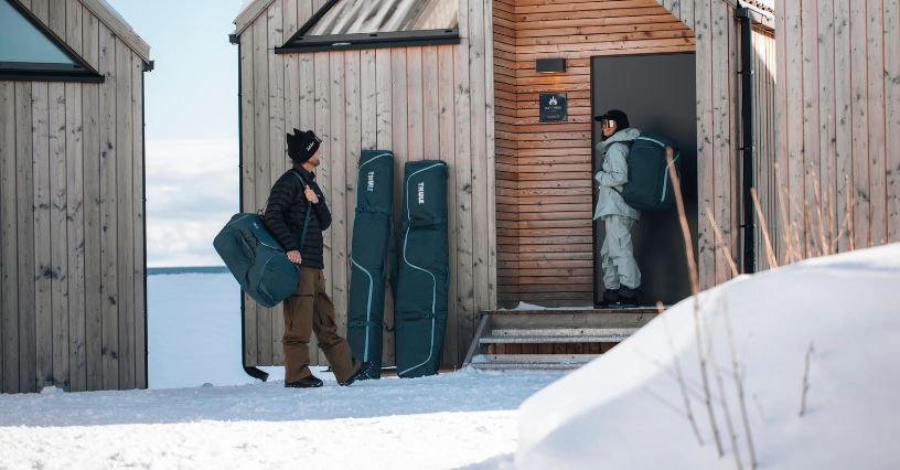 Kobieta i mężczyzna z dużymi torbami na buty narciarskie i pokrowcami na narty czekający przed drewnianym domkiem
