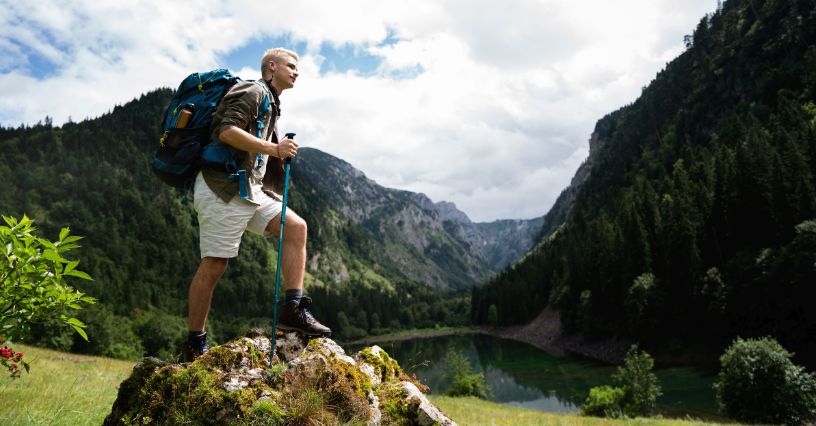 Mężczyzna z plecakiem i kijkami trekkingowymi stojący na skale na tle gór