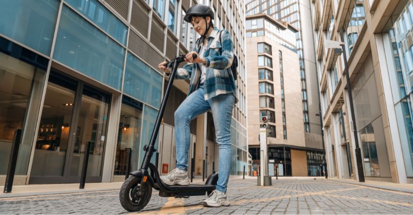 Kobieta w kasku stojąca na hulajnodze elektrycznej na środku ulicy między budynkami