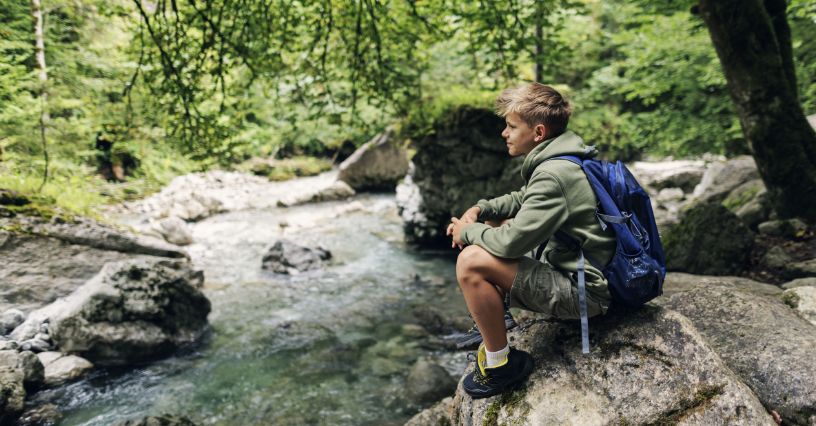 Nastolatek z plecakiem odpoczywający na skale nad rzeką w czasie wyprawy górskiej