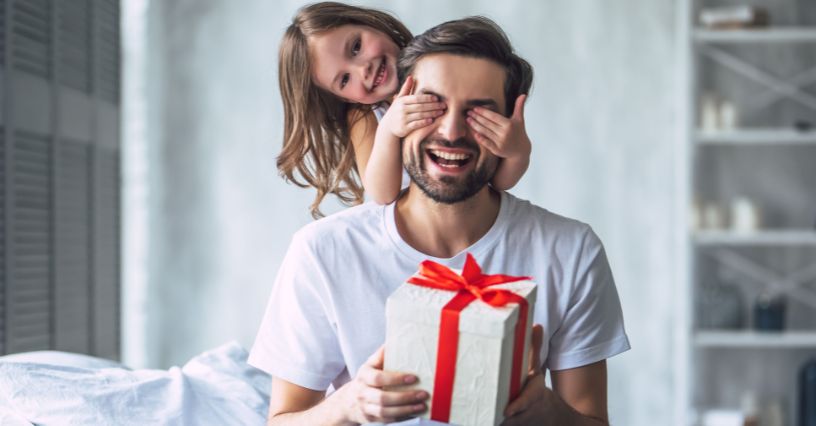 Dziewczynka zakrywająca oczy ojcu trzymającemu pudełko z prezentem