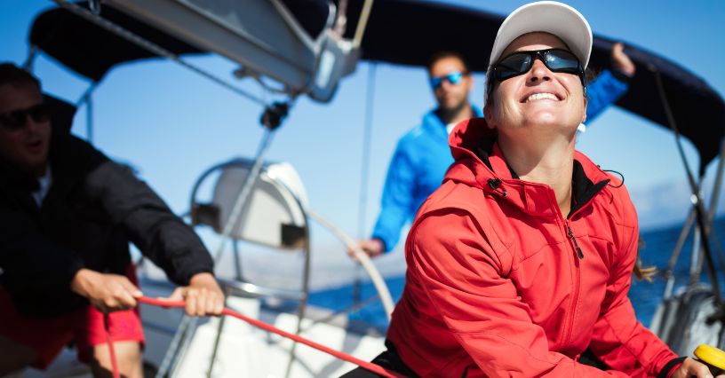 Uśmiechnięta kobieta w okularach przeciwsłonecznych i kurtce żeglarskiej patrząca w niebo w czasie żeglowania