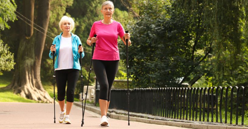 Dwie starsze kobiety spacerujące parkowymi ścieżkami z kijkami do nordic walking
