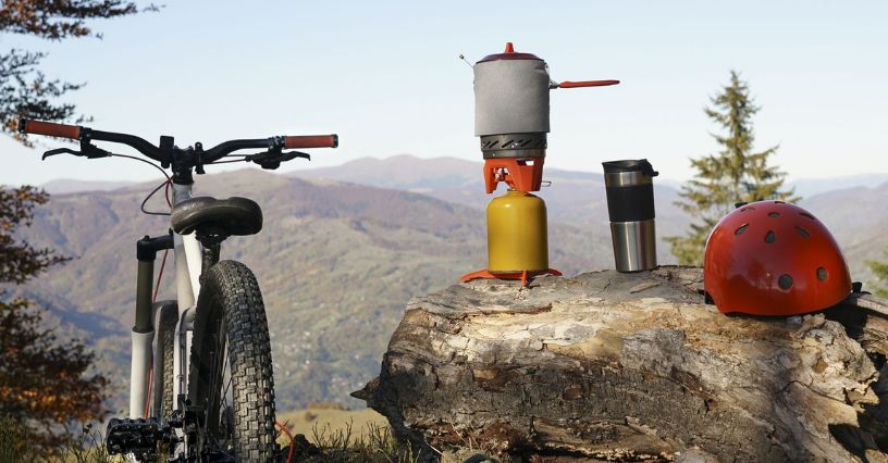 Kuchenka turystyczna na tle gór, obok roweru górskiego i czerwonego kasku rowerowego