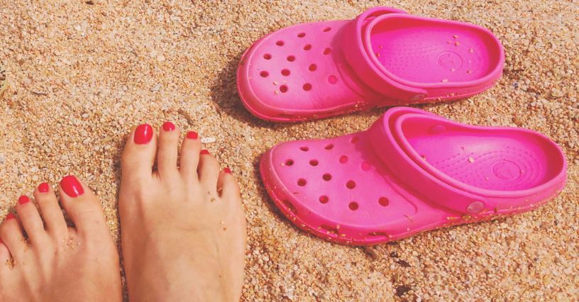 Klapki Crocs i kobiece stopy z paznokciami pomalowanymi na czerwono leżące na piasku