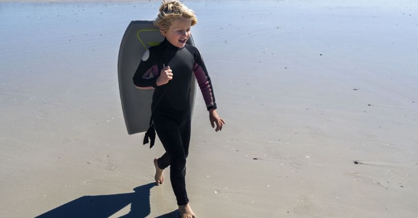 Chłopiec w długiej piance neoprenowej niosący na ramieniu deskę surfingową idący po plaży