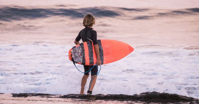 Chłopiec w krótkiej piance neoprenowej trzyma deskę surfingową stojąc na brzegu morza