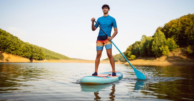 Mężczyzna stoi na desce SUP na spokojnym jeziorze, otoczony zielonymi wzgórzami