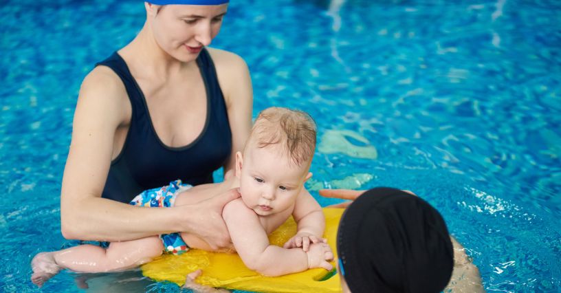 Mały chłopczyk w pieluszce na basen leży na desce do nauki pływania pod opieką instruktorki
