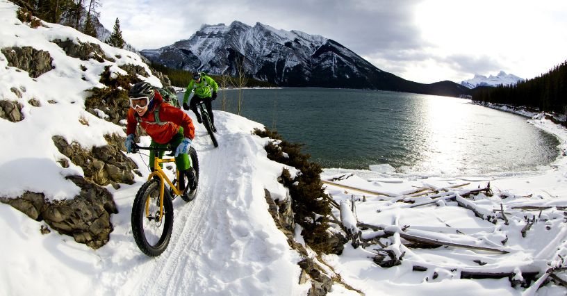 Mężczyzna na fatbike'u jedzie pewnie po zaśnieżonym szlaku koło stawu w górach