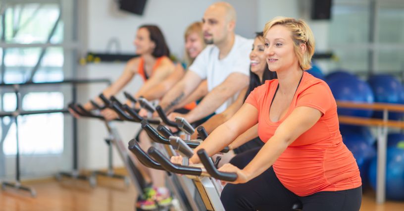 Kobieta w trzecim trymestrze ciąży ćwiczy na rowerze stacjonarnym w grupie