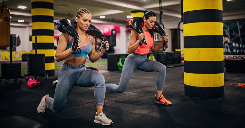 Dwie kobiety wykonują wykroki z workiem na barkach w klubie fitness