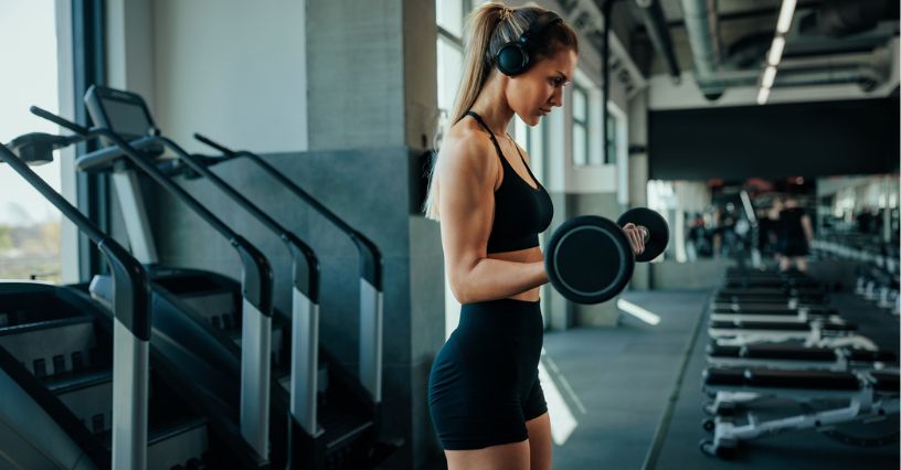 Kobieta w słuchawkach ćwicząca biceps z gryfem prostym na siłowni