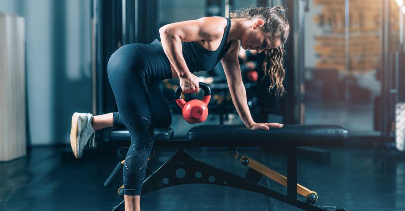 Kobieta ćwiczy wiosłowanie jednorącz z kettlebellem na ławce treningowej w siłowni