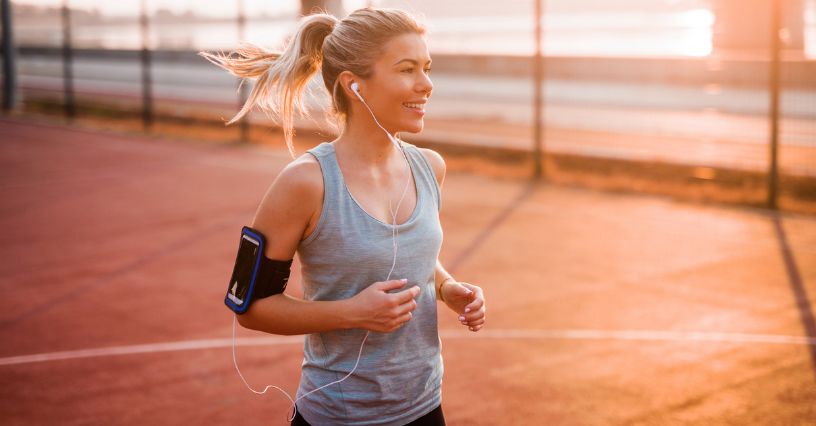 Kobieta w stroju sportowym biega o poranku słuchając muzyki puszczanej z telefonu schowanego w etui na ramieniu