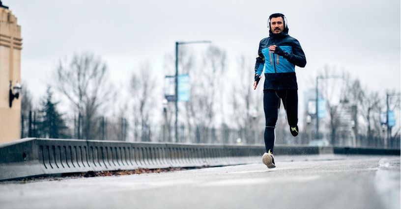 Biegacz w kurtce przeciwdeszczowej i ze słuchawkami biegnie miejską ulicą w deszczowy dzień