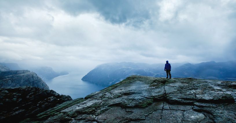Mężczyzna w kurtce przeciwdeszczowej stoi na szczycie góry obserwując górski widok w czasie nadchodzącego deszczu