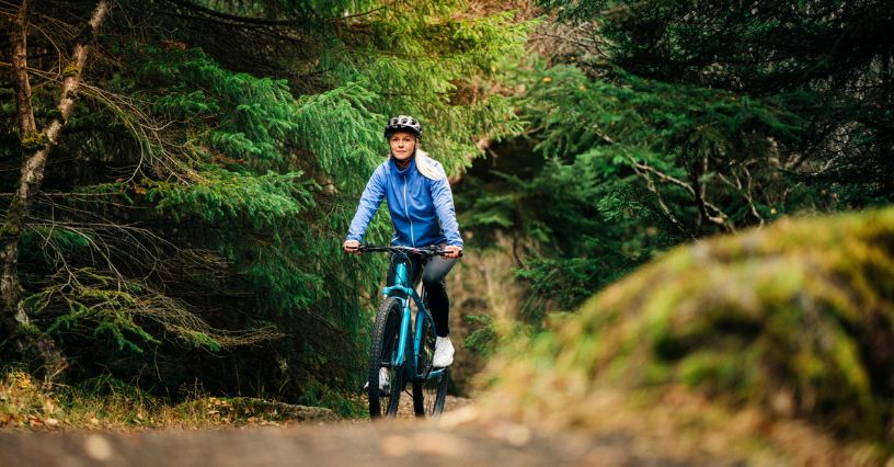 Kobieta w kasku i rowerowej kurtce jedzie leśną ścieżką na rowerze MTB