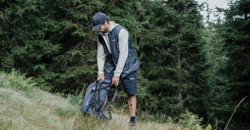 Mężczyzna w odzieży turystycznej zapina plecak Jack Wolfskin podczas postoju na górskiej polanie