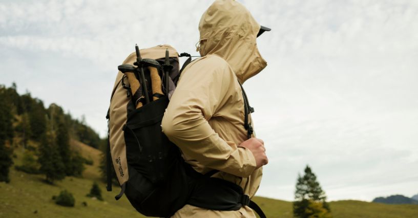 Mężczyzna w kurtce przeciwdeszczowej z kapturem maszeruje górską polaną z plecakiem turystycznym Jack Wolfskin