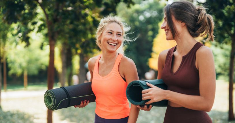 Dwie kobiety w strojach treningowych i z matami do ćwiczeń idą parkiem na zajęcia pilates